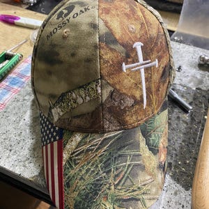 May include: A camouflage baseball cap with the Mossy Oak logo. The cap features an American flag design on the brim and a white embroidered cross on the side. The cap is made of a durable fabric.
