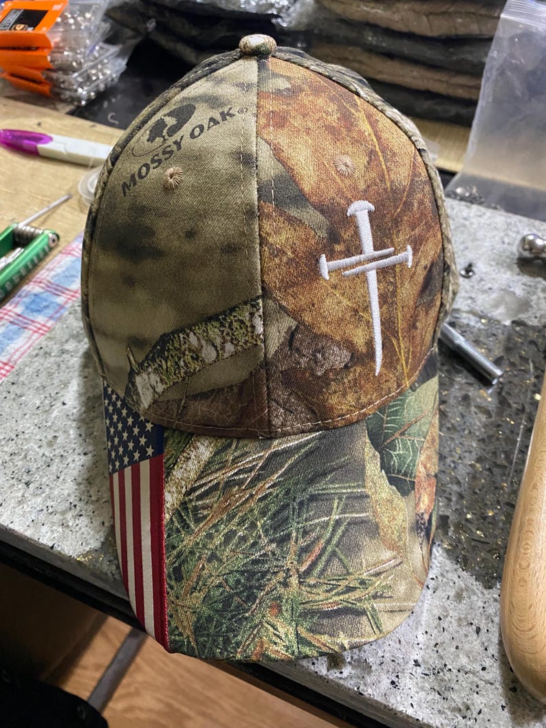 May include: A camouflage baseball cap with the Mossy Oak logo. The cap features an American flag design on the brim and a white embroidered cross on the side. The cap is made of a durable fabric.