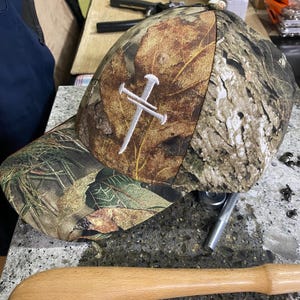 May include: Camouflage baseball cap with a white cross design. The cap features a leaf pattern in shades of brown, green, and tan. The cross is embroidered on the front. The cap has a curved brim and a button on top.