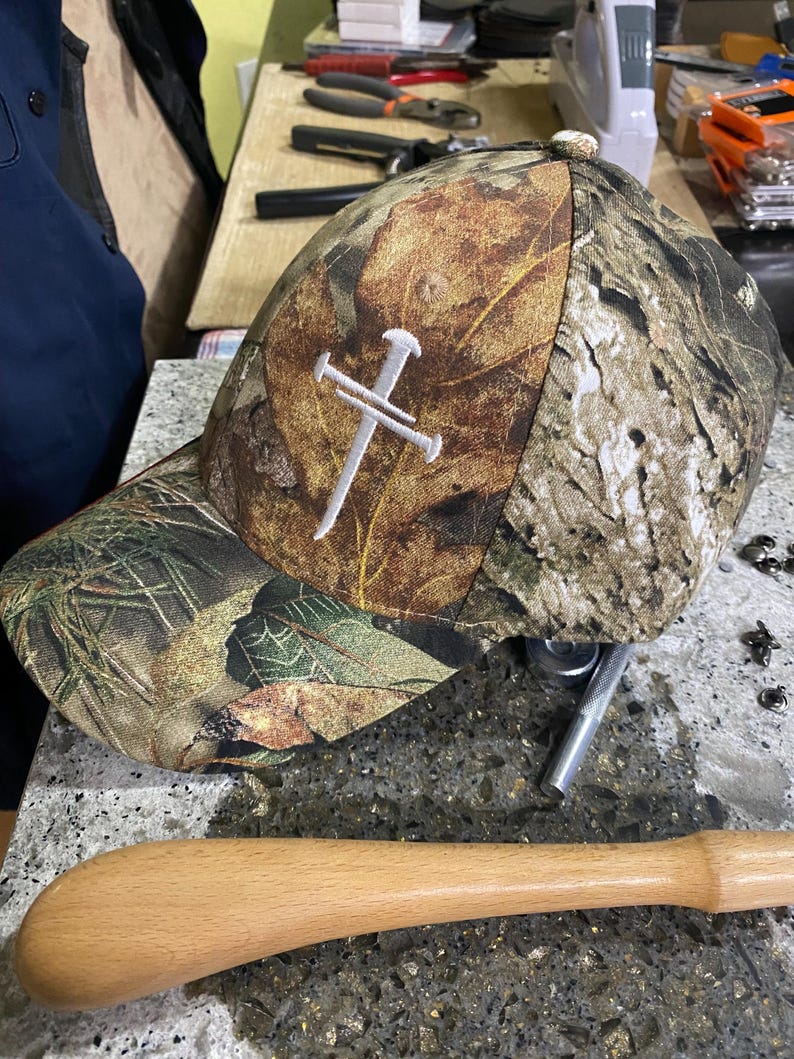 May include: Camouflage baseball cap with a white cross design. The cap features a leaf pattern in shades of brown, green, and tan. The cross is embroidered on the front. The cap has a curved brim and a button on top.