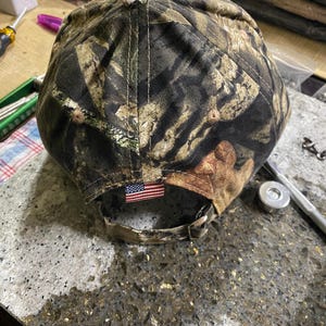 May include: A camouflage baseball cap with a curved brim. The cap features a brown, green, and tan pattern. An American flag patch is sewn on the back. The cap has an adjustable strap.