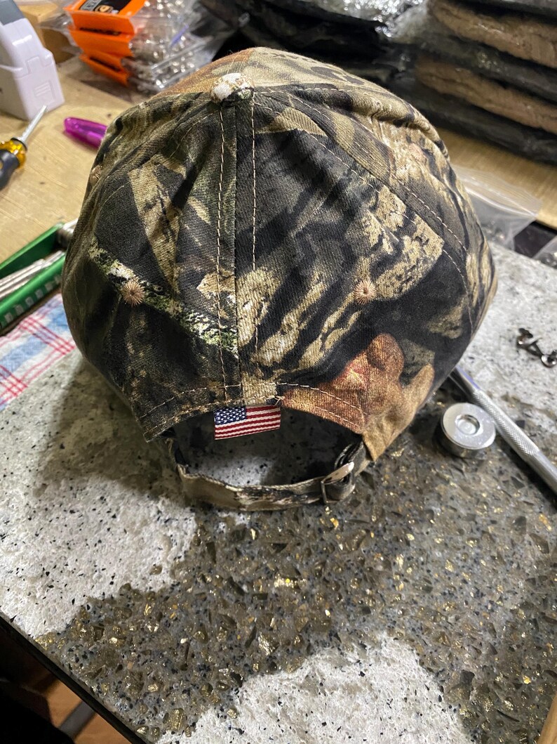 May include: A camouflage baseball cap with a curved brim. The cap features a brown, green, and tan pattern. An American flag patch is sewn on the back. The cap has an adjustable strap.