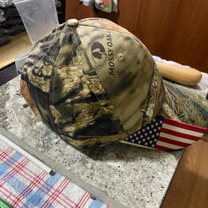 May include: A camouflage baseball cap with the text "Mossy Oak" and an American flag design on the brim. The cap features a brown, green, and black camouflage pattern.