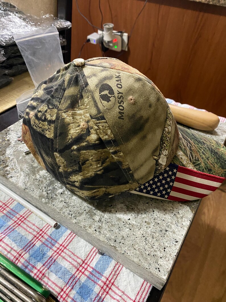 May include: A camouflage baseball cap with the text "Mossy Oak" and an American flag design on the brim. The cap features a brown, green, and black camouflage pattern.