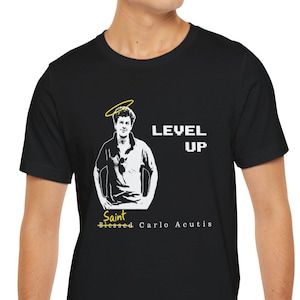 May include: Black t-shirt featuring a graphic of a man with a halo, the words "LEVEL UP", and "Saint Blessed Carlo Acutis". The design is in white and yellow. The shirt is worn by a person.
