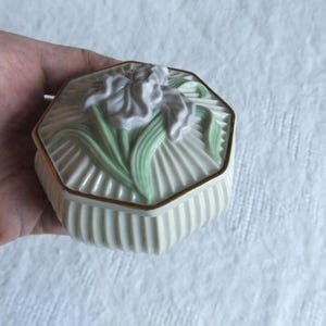 May include: A cream-colored, octagonal trinket box with a lid featuring a raised iris design. The iris has lavender petals and green leaves. The box has a ribbed body and gold trim. A decorative item.