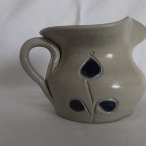 May include: A small, gray ceramic pitcher with a blue floral design. The pitcher has a handle and a wide spout.