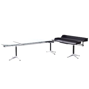 May include: A large glass and metal desk with a black metal extension. The desk has a curved front edge and a silver base with three legs. The extension has a black top with two shelves and a silver base with three legs.