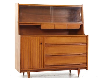 May include: A mid-century modern wooden buffet with a glass-front hutch. The buffet has a cabinet door with vertical grooves, three drawers, and a top shelf with two smaller drawers. The buffet is made of light wood and has a warm, natural finish.