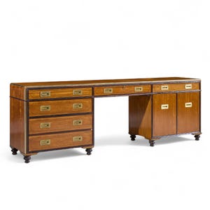 May include: A large, wooden desk with a warm brown finish. It features multiple drawers, cabinets, and a central open space for a chair. The desk has brass hardware and dark brown trim, and is supported by short, rounded legs.