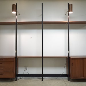 May include: A modular wall unit with black metal poles and brown wood shelves. The unit includes a desk with drawers and a cabinet with sliding doors.