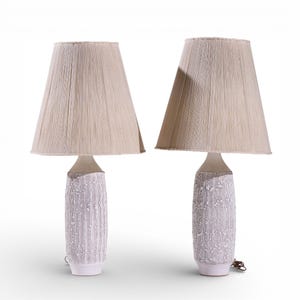 May include: Two matching table lamps with beige fabric shades. The lamps have a textured, white ceramic base with a cylindrical shape. The shades are a light beige color, and the lamps are set against a white background.
