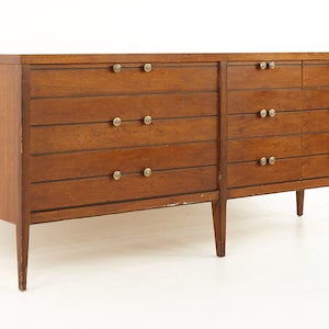 May include: A long, wooden dresser with six drawers and brass knobs. The dresser is made of a dark wood and has a simple, mid-century modern design.