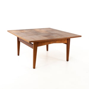 May include: A square wooden coffee table with a light brown finish and four legs. The table has a simple, mid-century modern design.