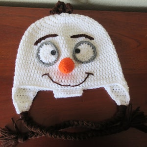 May include: White crocheted snowman hat with brown yarn for hair and a long brown yarn tassel. The hat has a stitched-on orange nose, gray eyes, and a brown stitched-on smile.
