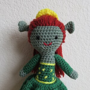 May include: A green crocheted doll with red hair and a yellow crown. The doll is wearing a green dress with yellow accents.