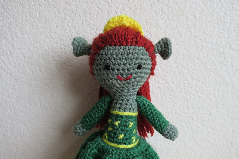 May include: A green crocheted doll with red hair and a yellow crown. The doll is wearing a green dress with yellow accents.