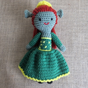 May include: A crocheted doll wearing a green and yellow dress with a yellow crown. The doll has grey facial features with black eyes and a red mouth.