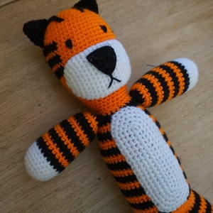 May include: A handmade crochet tiger toy in orange, black, and white. The tiger has black stripes, a white face and belly, and black ears. The arms and legs are outstretched, giving the toy a playful appearance.