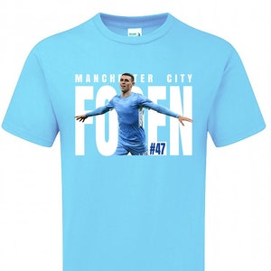 May include: Light blue t-shirt with a white graphic of a football player celebrating a goal. The graphic includes the text "MANCHESTER CITY" and "FODEN #47".