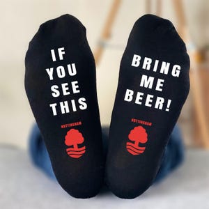 May include: Black socks with white text that reads "IF YOU SEE THIS" on one sock and "BRING ME BEER!" on the other. Each sock has a red tree logo and the word "NOTTINGHAM" below the text.