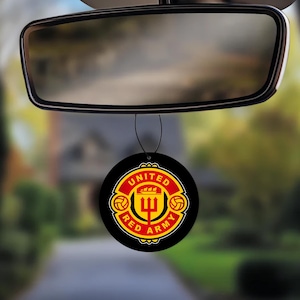 Op de afbeelding: Een zwarte, ronde auto luchtverfrisser met de tekst "UNITED RED ARMY" in rood en geel. Het ontwerp toont een wapen met een drietand en voetballen, hangend aan de achteruitkijkspiegel.