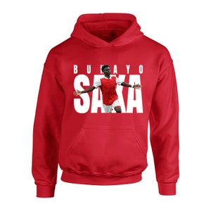 May include: A red hoodie featuring the name "BUKAYO SAKA" in white block letters above a photo of a soccer player in a red and white jersey. The hoodie has a front pocket and a hood.