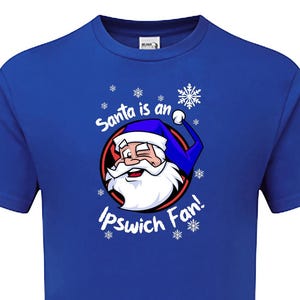May include: A royal blue t-shirt with a white graphic of Santa Claus winking and wearing a blue hat. The text "Santa is an Ipswich Fan!" is printed in white below the graphic.