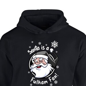 May include: Black hooded sweatshirt with a white graphic of Santa Claus wearing a black hat and a white beard. The graphic says "Santa is a Fulham Fan!" with snowflakes around the graphic.