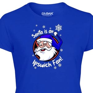 May include: Royal blue t-shirt featuring a cartoon Santa Claus with a blue hat and white beard. The text "Santa is an Ipswich Fan!" is printed on the shirt, surrounded by snowflakes. Gildan Performance label is visible.