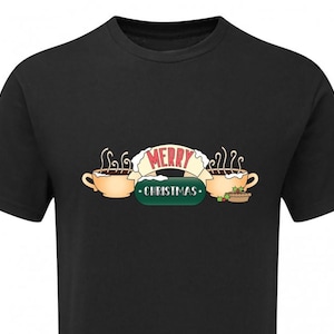 Op de afbeelding: Zwart T-shirt met een feestelijke afbeelding. Het ontwerp toont twee beige mokken warme chocolademelk met stoom, een banner met de tekst "MERRY CHRISTMAS" en een groene ovaal met dezelfde tekst. Rechts staat een kleine taart.