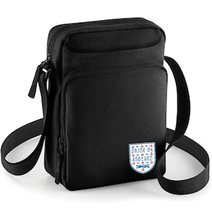 May include: Black crossbody bag with a rectangular shape and a long adjustable strap. The bag features a front pocket and a zipper closure. A patch with the text "Pride of England" and a crest is on the front.