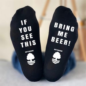 May include: Black socks with white text that reads "IF YOU SEE THIS" on one sock and "BRING ME BEER!" on the other. The socks also feature a white tree logo and the word "NOTTINGHAM".