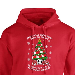 May include: Red hooded sweatshirt with a white Christmas tree design. The tree is decorated with soccer balls and scarves. The text reads "Jingle Bells Jingle Bells Jingle All The Way" and "Oh, What Fun It Is To See Nottingham Win Away".