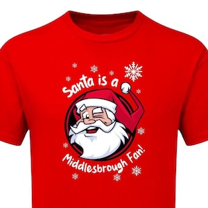Könnte beinhalten: Rotes T-Shirt mit einem Cartoon-Weihnachtsmann, der zwinkert und einen roten Hut und einen weißen Bart trägt, in einem schwarzen Kreis. Der Text "Santa is a Middlesbrough Fan!" steht über und unter der Weihnachtsmann-Illustration, mit Schneeflocken.