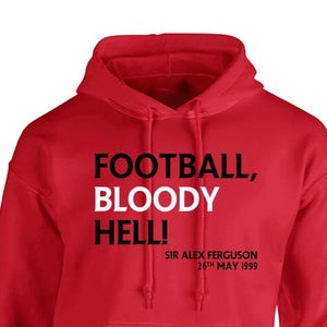 May include: A red hooded sweatshirt with the text "FOOTBALL, BLOODY HELL!" in black and white. Below the text is "SIR ALEX FERGUSON 26th MAY 1999" in black. The sweatshirt has a drawstring hood.