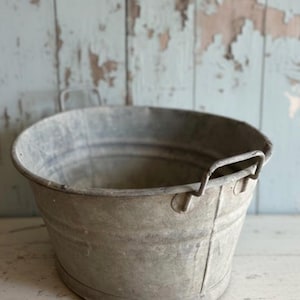 Op de afbeelding: Een ovale metalen emmer met twee kleine handvatten, met tekenen van ouderdom en slijtage. De emmer heeft een verweerde uitstraling. De achtergrond is een lichtblauwe houten muur met afbladderende verf.