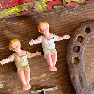 Op de afbeelding: Twee kleine, beschilderde beeldjes van kinderen met blond haar en roze wangen. De beeldjes dragen eenvoudige, witte jurken en staan ​​met hun armen uitgestrekt. De beeldjes staan ​​op een houten oppervlak met een roestig hoefijzer en een zilveren ketting.