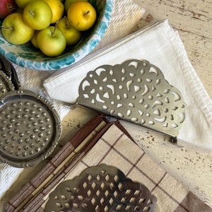 Op de afbeelding: Een stilleven met een decoratieve metalen servethouder met een uitgesneden ontwerp, met gevouwen bruin-wit geruite servetten. Een kom met groene en gele appels staat in de buurt, samen met een witte servet en een zilveren onderzetter.