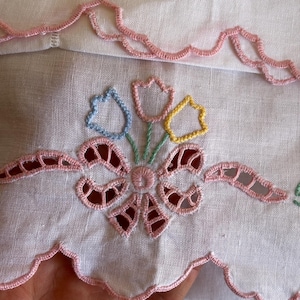 Puede incluir: Toalla de té de lino blanco con bordes festoneados rosas y flores bordadas en azul, amarillo y verde. Las flores están centradas alrededor de un lazo rosa.