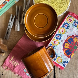Könnte beinhalten: Eine braune Keramiktasse mit Henkel, eine braune Keramiksaucers und ein brauner Keramikteller. Die Tasse, die Untertasse und der Teller befinden sich auf einer Holzoberfläche mit einem rosa Stoff, auf dem das Wort "bar" gestickt ist.