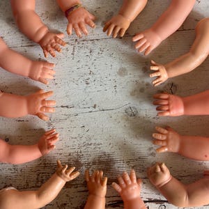 May include: A collection of ten doll arms, made of a light pink plastic, arranged in a circle on a white wooden surface. The arms are all bent at the elbow, with the hands facing inwards towards the center of the circle.