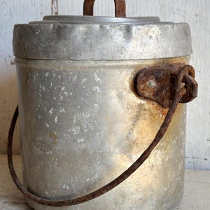 Puede incluir: Un cubo de metal vintage con tapa y asa. El cubo es plateado con un asa oxidada y una bisagra oxidada. El cubo tiene forma cilíndrica y probablemente se utiliza para transportar agua u otros líquidos.