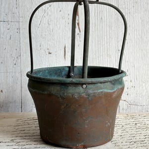 May include: A vintage copper bucket with a dark patina and a metal handle. The bucket has a rounded base and a slightly flared rim. The handle is arched and attached to the bucket with metal loops. The copper has a mix of brown and green hues.