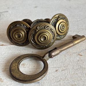 Könnte beinhalten: Drei antike Messing-Schrankknöpfe mit verzierten Designs und einem passenden Messing-Skelettschlüssel.