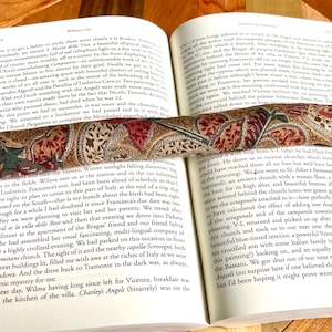 May include: An open book with a patterned bookmark resting between the pages. The bookmark has a paisley design in red, brown, and green. The book's pages are yellowed, and text is visible.