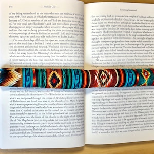 May include: A patterned bookmark lies on an open book. The bookmark has a geometric design in blue, orange, and brown. The book's pages are visible, with text. The bookmark is approximately 20 cm long.
