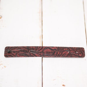 May include: A brown leather bracelet with a floral pattern. The bracelet has a metal snap closure.