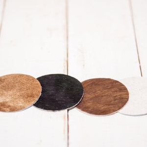 May include: Four round coasters made of different materials. The coasters are brown, black, brown, and white. The coasters are arranged in a row on a white wooden surface.