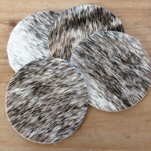 May include: Four round cowhide coasters in shades of white and brown. The coasters are made of natural cowhide and have a soft, fuzzy texture.